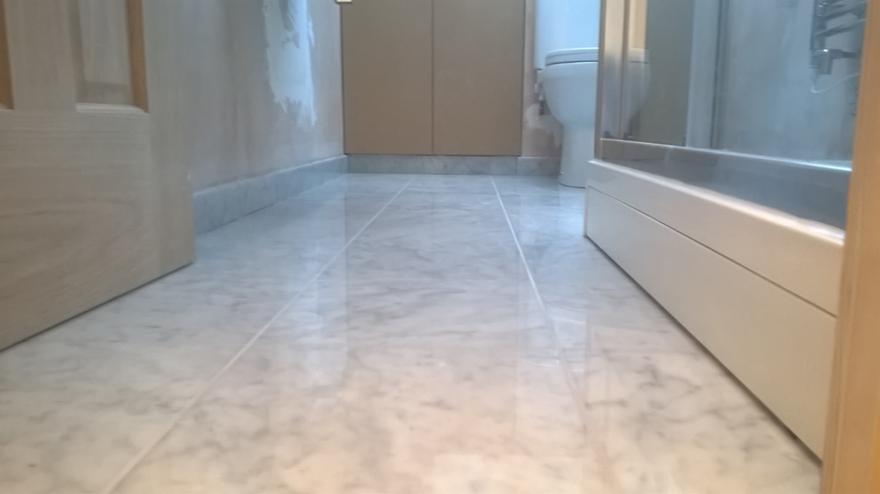 Jasna, marmurowa posadzka w łazience z widoczną kabiną prysznicową i fragmentem ściany z nałożoną gładzią. Widok z niskiej perspektywy.