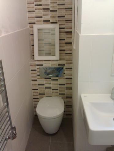 Nowoczesna toaleta podwieszana w małej łazience z mozaiką w odcieniach brązu i beżu. Biała umywalka i rama nad spłuczką. Szare płytki podłogowe.