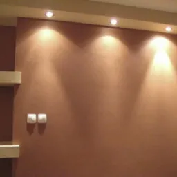 Czekoladowa ściana z dekoracyjnym tynkiem, oświetlona trzema punktowymi lampami sufitowymi. Z lewej strony dwie półki i włączniki światła.