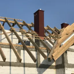 Wznoszenie konstrukcji drewnianej dachu na budynku z białych bloczków, widoczne kominy w kolorze bordowym, jasne niebo w tle.