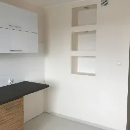 Wnętrze kuchni z białymi szafkami, ciemnym blatem, drewnianymi frontami szafek dolnych i wnękami w ścianie pomalowanej na kremowo.