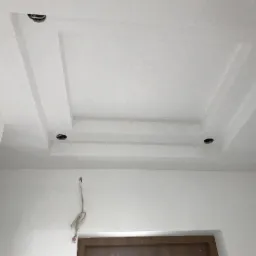 Biały sufit z dekoracyjnym, kaskadowym obniżeniem i otworami na oświetlenie punktowe, widoczne kable zwisające ze ściany, surowy stan wykończenia.