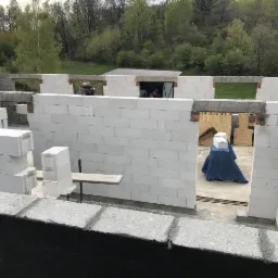 Wznoszenie ścian domu z białych bloczków, widoczne otwory okienne i drzwiowe, zabezpieczone materiały budowlane wewnątrz budynku, krajobraz zieleni w tle.