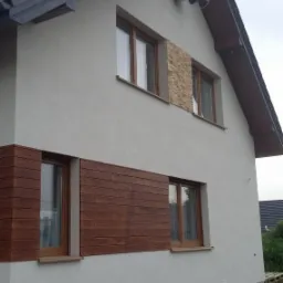 Wykończona elewacja domu jednorodzinnego z elementami dekoracyjnymi z drewna i kamienia, widok z boku na tle zachmurzonego nieba.