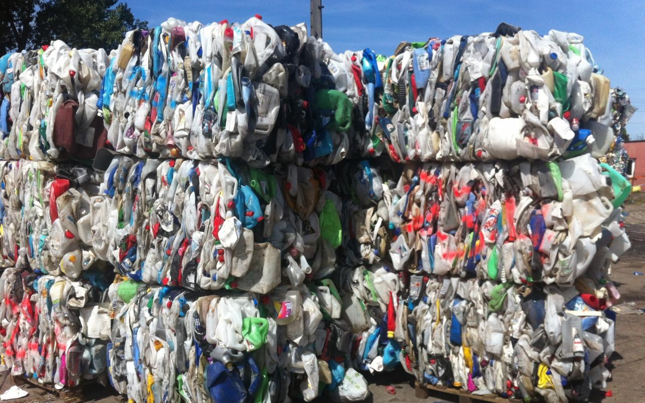 Stos sprasowanych, kolorowych plastikowych butelek i kanistrów, tworzących regularny blok na zewnątrz, w słoneczny dzień. Widoczne elementy etykiet.