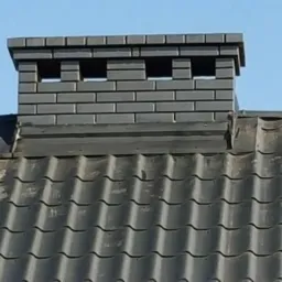 Szary komin z cegły klinkierowej z otworami wentylacyjnymi na dachu pokrytym ciemną dachówką ceramiczną. Widok z dołu na tle błękitnego nieba.