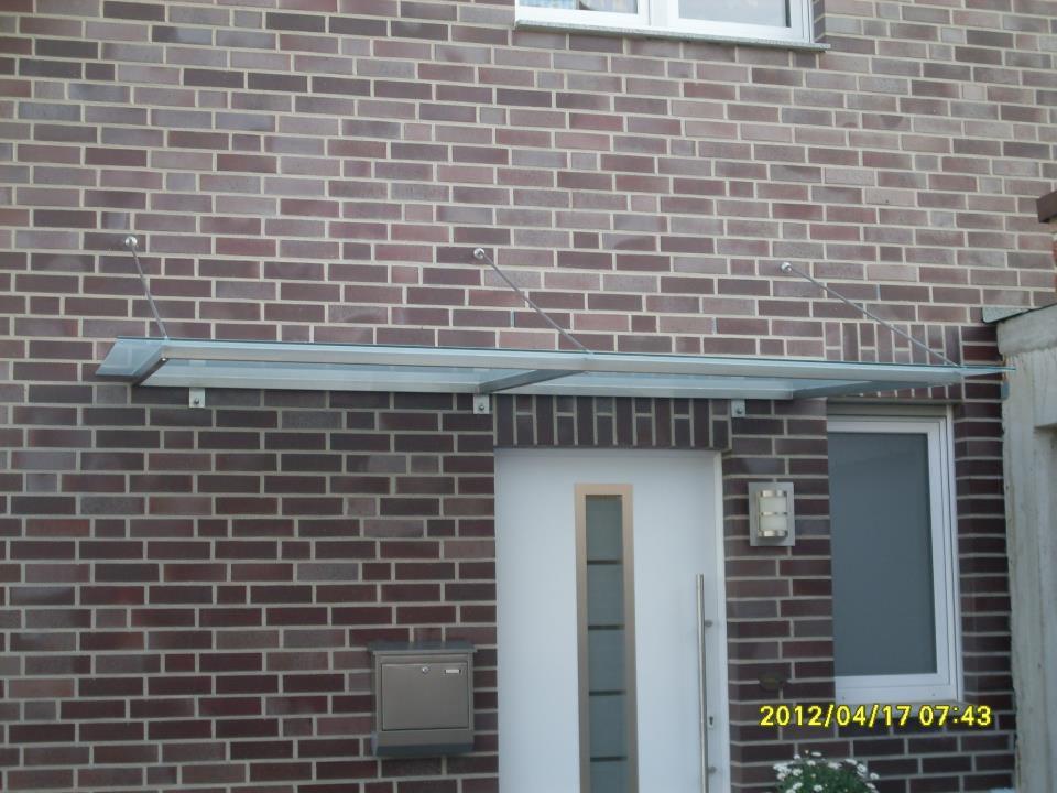 Szklany daszek nad wejściem do domu z ceglaną elewacją, wsparty na metalowych linkach, widoczna skrzynka na listy i lampa zewnętrzna.