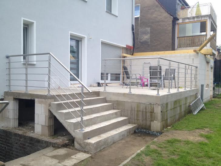 Zewnętrzne schody betonowe prowadzące na taras z balustradą nierdzewną z poziomymi prętami, widoczne niedokończone prace budowlane i surowe wykończenie.