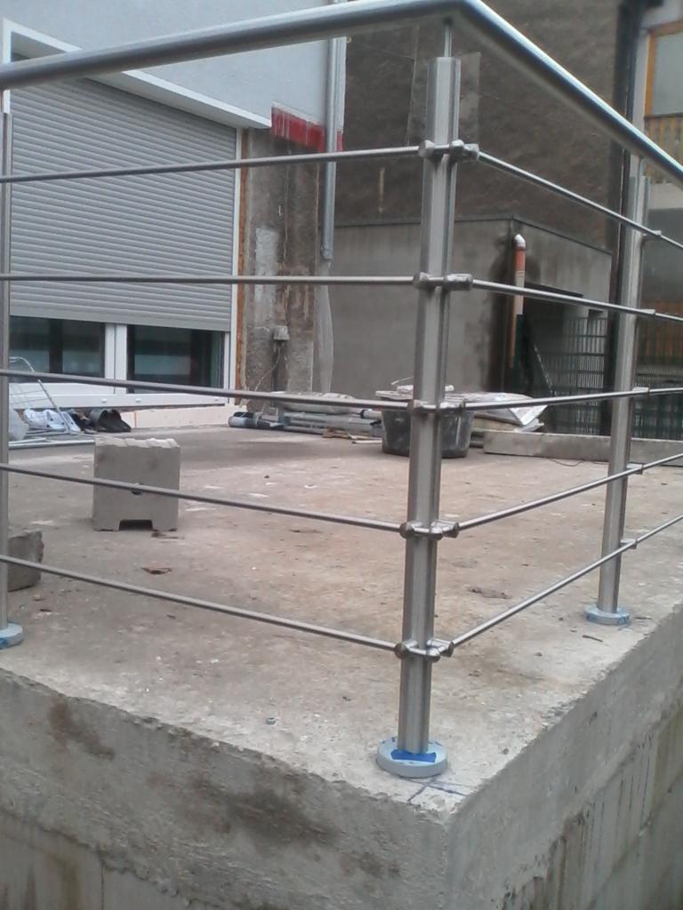 Montaż balustrady z rurek na betonowym balkonie, widoczne narzędzia i materiały budowlane w tle.