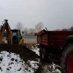Żółta koparka CAT nabiera ziemię na śnieżnym terenie, obok czerwona przyczepa i fragment zielonego traktora Fendt. W tle mur z bloczków.