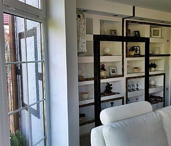 Wnętrze pokoju z białymi regałami w zabudowie, widoczne przez okno z białymi ramami, w tle fragment białej sofy.