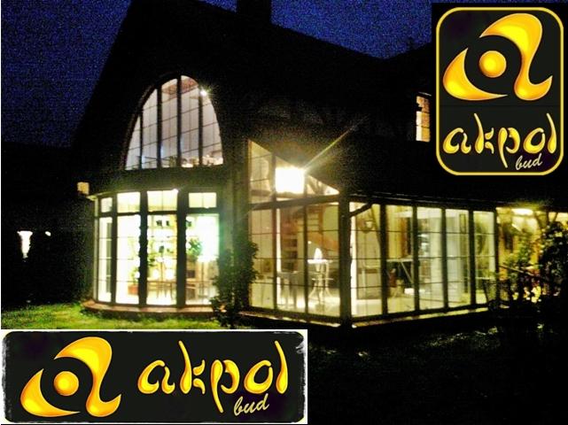 Oświetlony budynek z dużymi oknami w nocy, z logo firmy Akpol Bud na pierwszym planie i w rogu. Widoczne wnętrze i elementy konstrukcyjne.