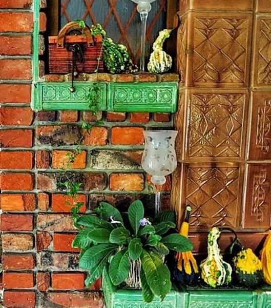 Dekoracyjna ściana z czerwonej cegły w Tychach, z zielonymi półkami eksponującymi dynie ozdobne, roślinę doniczkową i szklane świeczniki, obok ściana wyłożona brązowymi, wzorzystymi kaflami.