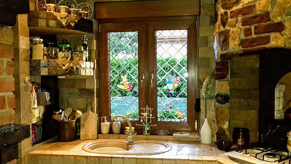 Rustykalna kuchnia z oknem z brązową ramą, ozdobionym naklejkami z motywem wielkanocnym, widok na zielony ogród, kamienne wykończenie ściany i półki z dekoracyjnymi naczyniami.