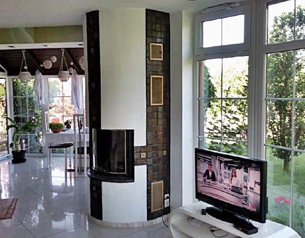 Jasne wnętrze z dużymi oknami PCV, nowoczesnym kominkiem obłożonym mozaiką i widokiem na zieleń za oknem.