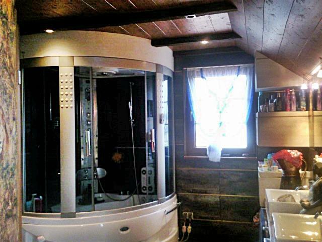 Kabina prysznicowa z hydromasażem w łazience z drewnianym sufitem i skośnym dachem, widok na okno z niebieską firanką i umywalki.