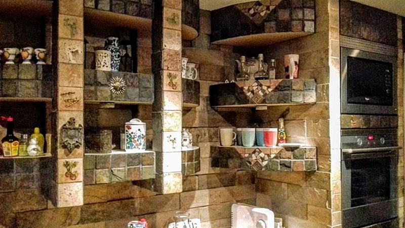 Wykończenie ściany w kuchni z nieregularnych kamiennych płytek, tworzących półki z dekoracjami i naczyniami, obok zabudowanego piekarnika z mikrofalówką.