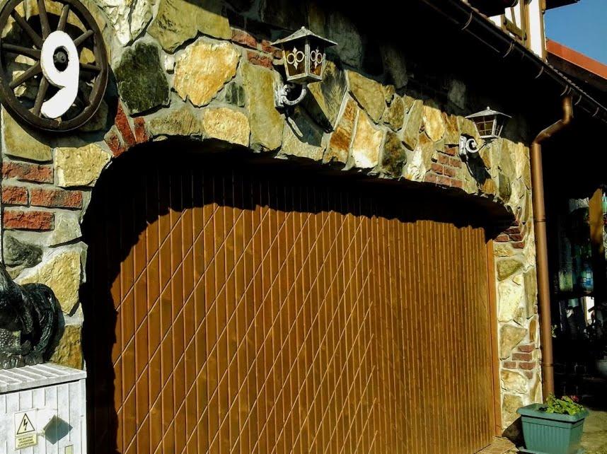 Drewniana brama garażowa z ukośnymi panelami, wkomponowana w kamienną elewację z ceglanymi akcentami, oświetlona dwoma stylizowanymi lampionami i ozdobiona dekoracyjnym kołem z numerem 9.