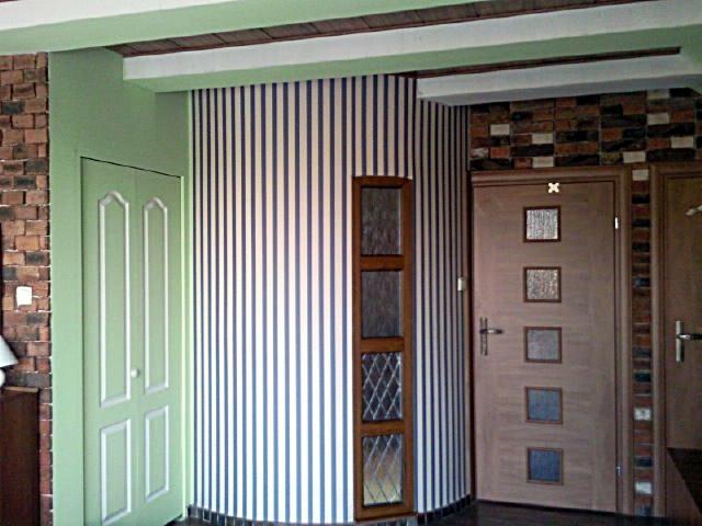 Wnętrze z zakrzywioną ścianą w biało-granatowe pasy, drzwi wewnętrzne z szybkami w drewnianej ramie, zielone drzwi dwuskrzydłowe i fragment ściany z cegły.