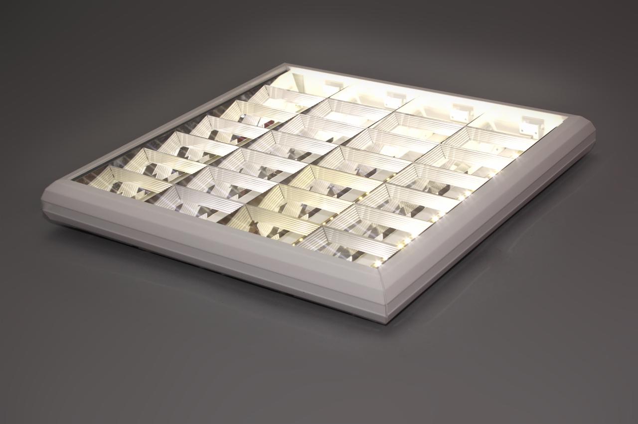 Kwadratowa lampa sufitowa z rastrem w białej obudowie, oświetlająca szarą powierzchnię. Widoczna struktura rastra i delikatne, ciepłe światło.