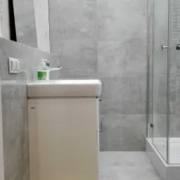 Wykończona łazienka w odcieniach szarości z umywalką nablatową na białej szafce, kabiną prysznicową ze szklanymi drzwiami i owalnym lustrem na ścianie pokrytej jasnymi płytkami.