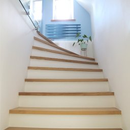 PracowniaARD - Białe schody z drewnianymi stopniami prowadzące do góry, z przeszkloną balustradą, niebieską ścianą z grzejnikiem i rośliną w tle.