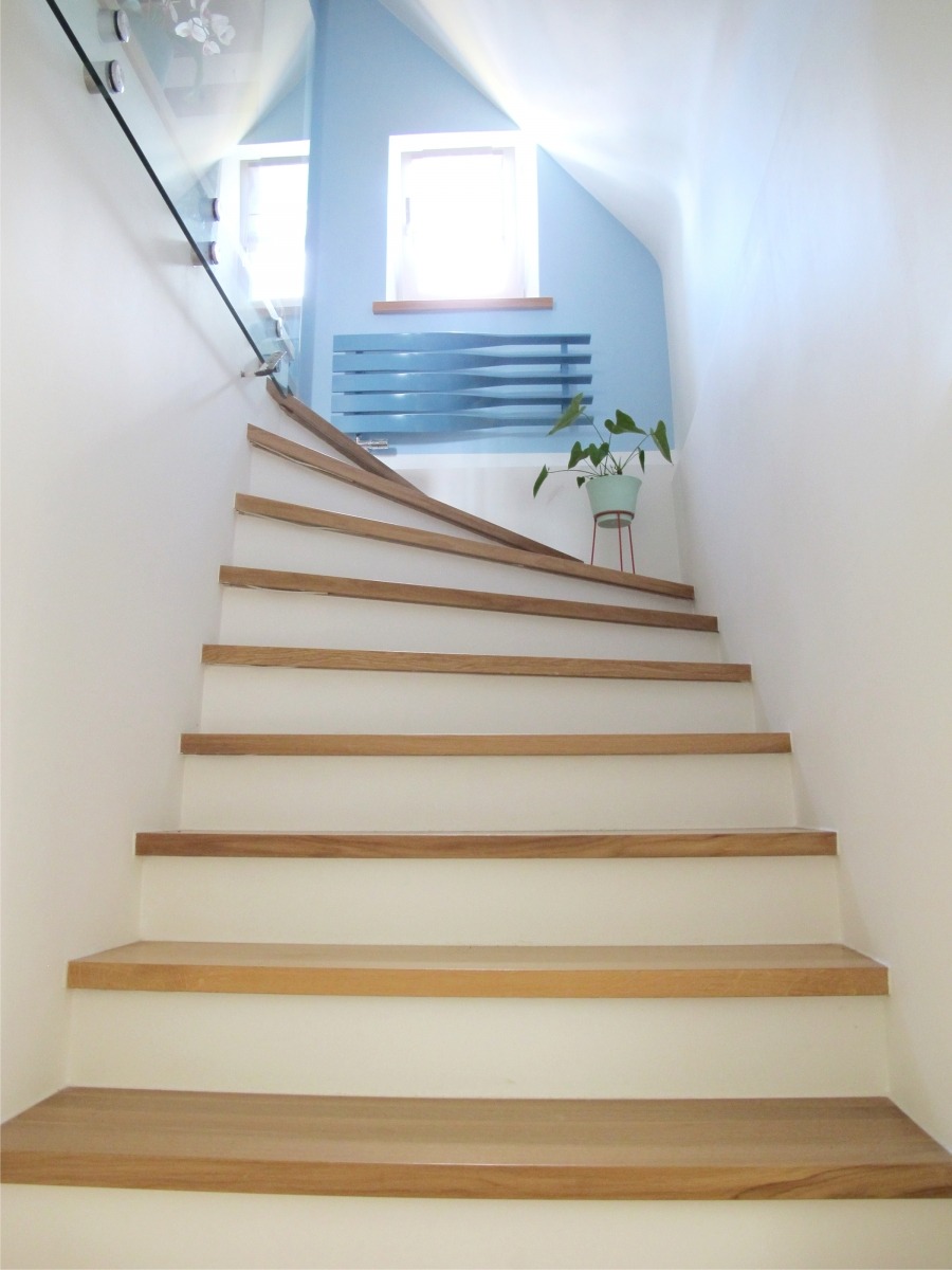 Białe schody z drewnianymi stopniami prowadzące do góry, z przeszkloną balustradą, niebieską ścianą z grzejnikiem i rośliną w tle.
