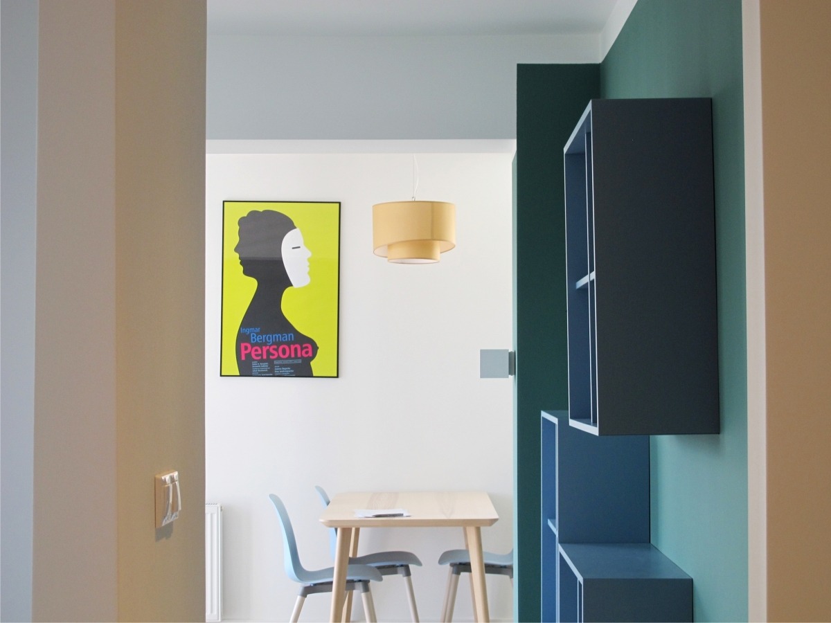 Nowoczesne wnętrze jadalni z plakatem Bergman Persona, drewnianym stołem, niebieskimi krzesłami i wiszącą lampą z abażurem, widok przez przejście.
