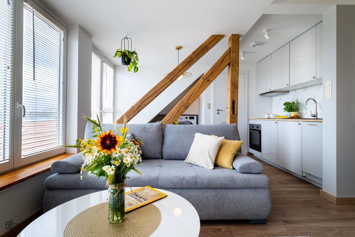 Jasne wnętrze mieszkania z szarą sofą, bukietem kwiatów na okrągłym stoliku, drewnianymi belkami konstrukcyjnymi oraz aneksem kuchennym w tle.