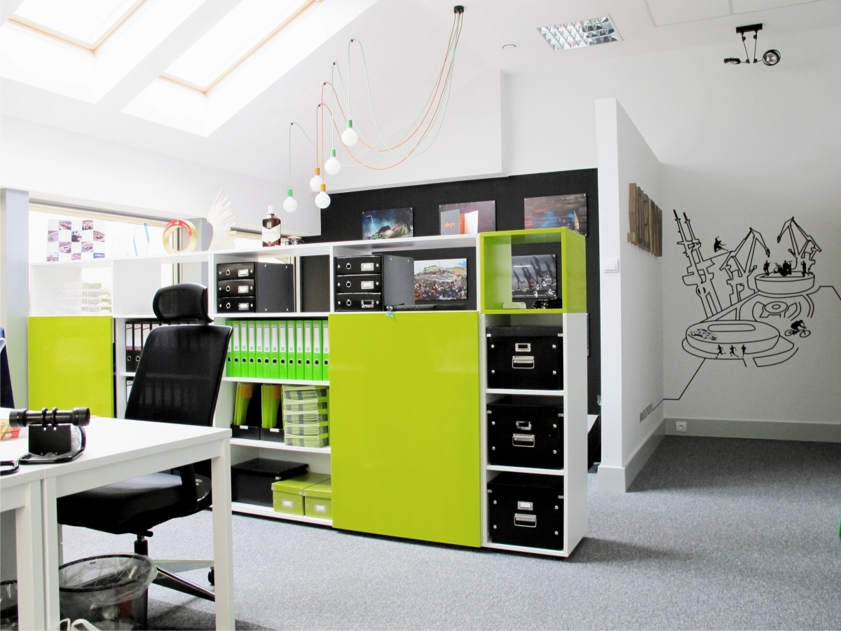 Nowoczesne biuro z białymi ścianami, czarną ścianą akcentującą, regałem z zielonymi akcentami i grafiką na ścianie. Biurko z czarnym krzesłem i widoczne segregatory.