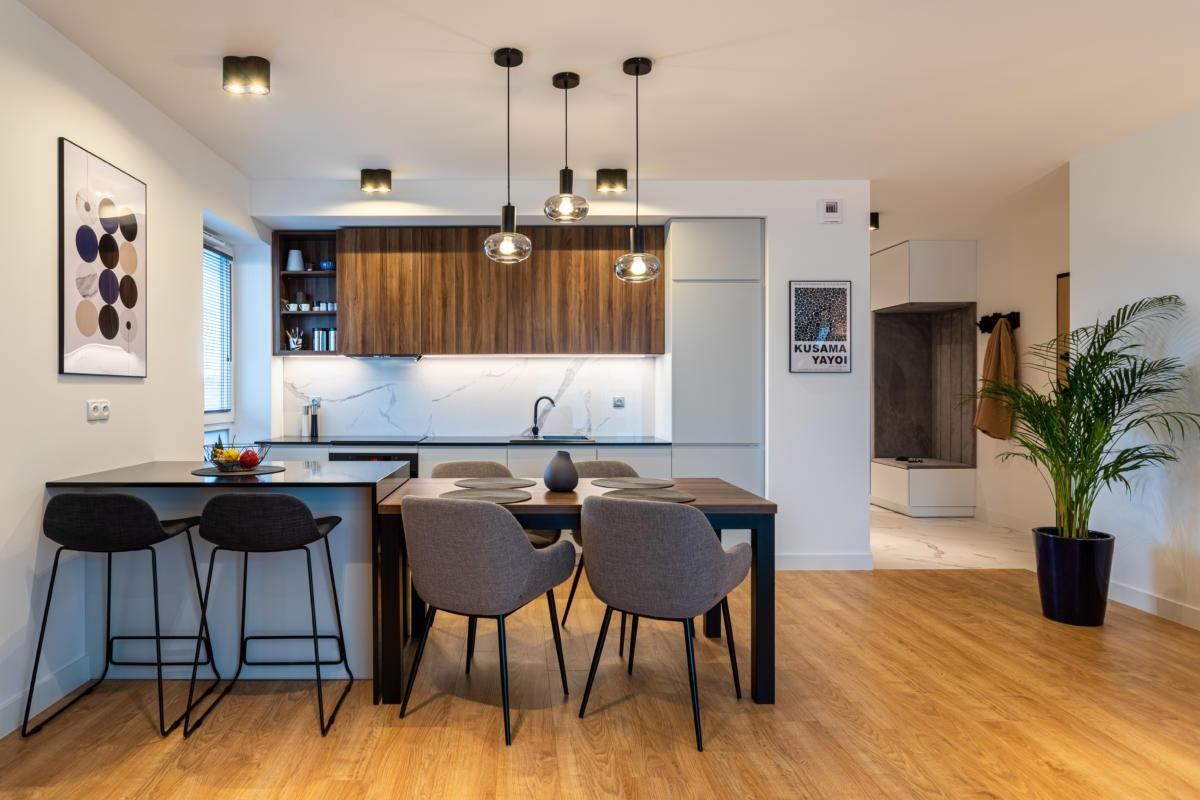 Nowoczesne wnętrze mieszkania z drewnianą podłogą, wyspą kuchenną z hokerami, stołem jadalnym i przeszklonymi lampami wiszącymi nad nim, oraz rośliną doniczkową w tle.