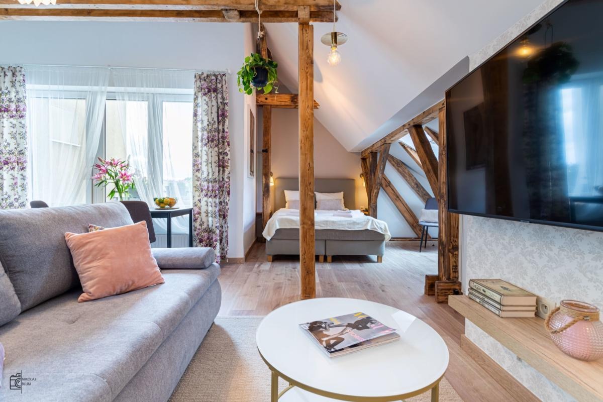 Jasne, przestronne wnętrze na poddaszu z drewnianymi belkami, szarą sofą z różową poduszką, okrągłym stolikiem z magazynem, łóżkiem w tle i telewizorem na ścianie w stylu skandynawskim.