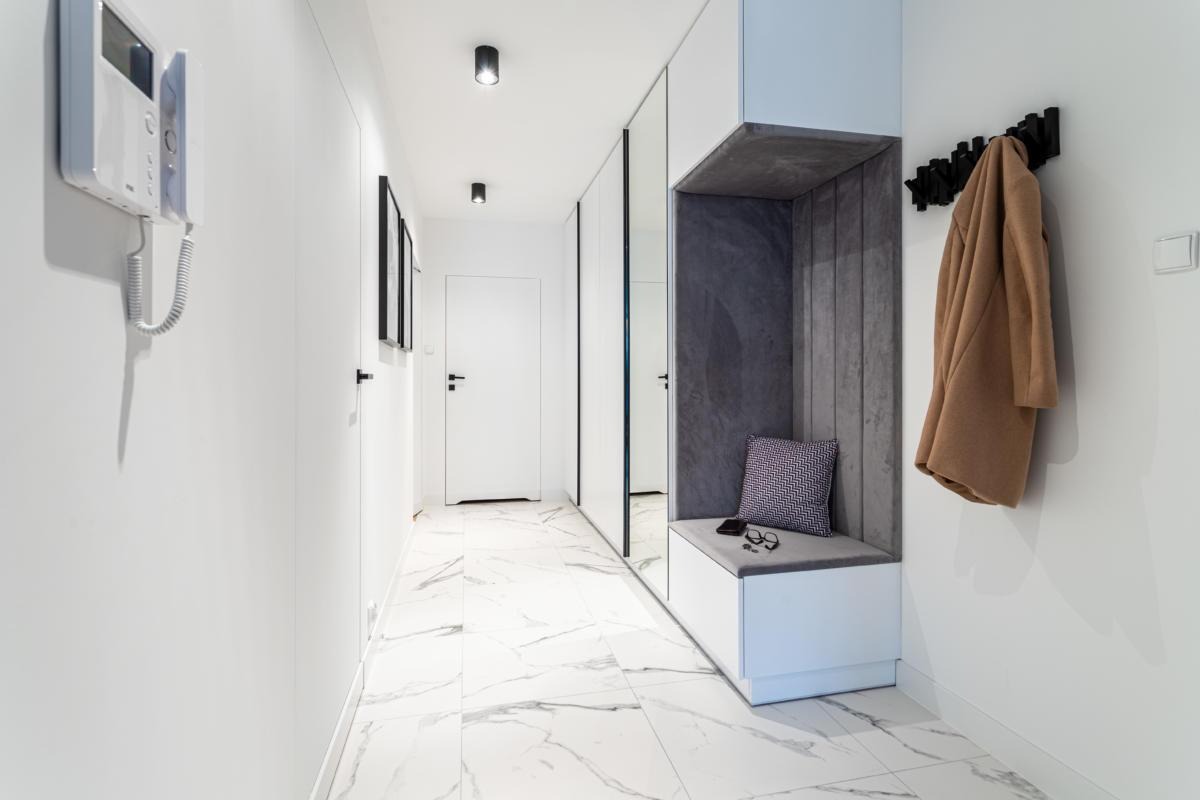 Nowoczesny przedpokój w bieli i szarości z marmurową podłogą, zabudowaną szafą z lustrem, siedziskiem z szarą tapicerką, poduszką w zygzaki, okularami, telefonem i beżowym płaszczem wiszącym...