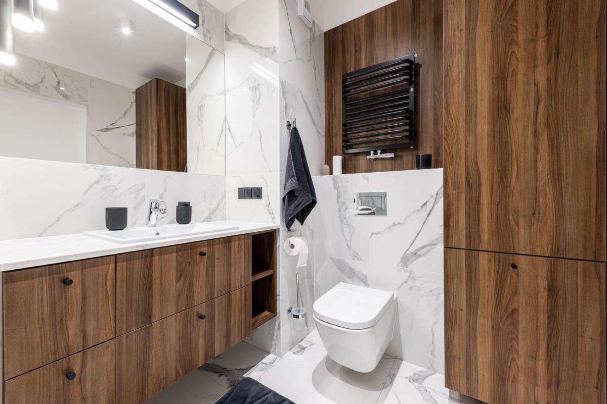 Nowoczesna łazienka z marmurowymi płytkami, drewnianymi szafkami pod umywalką, wiszącą toaletą i czarnym grzejnikiem drabinkowym.