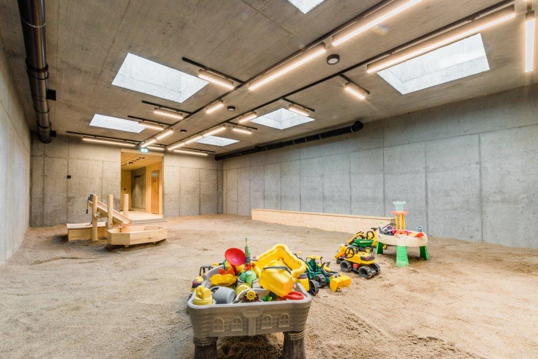 Betonowe wnętrze sali zabaw z piaskownicą, oświetlone lampami sufitowymi i świetlikami; widoczne zabawki i drewniane konstrukcje.