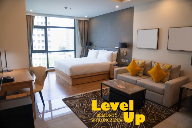 Wykończony apartament hotelowy z dużym oknem, łóżkiem, sofą z żółtymi poduszkami, biurkiem i dywanem w odcieniach brązu.