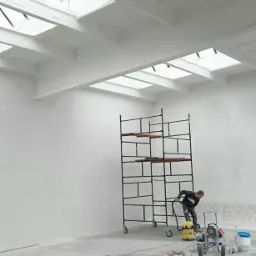 Wnętrze w trakcie prac wykończeniowych; widoczne rusztowanie, agregat malarski, odkurzacz przemysłowy i pracownik przygotowujący podłoże przed aplikacją farby.