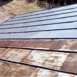 Częściowo odnowiony dach z metalowymi płytami, ukazujący kontrast między nową szarą powłoką a starą, zardzewiałą powierzchnią.