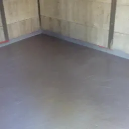 Szara, świeżo położona posadzka w rogu pomieszczenia z surowymi ścianami betonowymi i widocznymi łączeniami płyt.