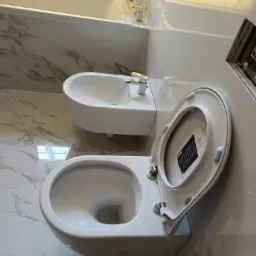 Biała toaleta i bidet na tle marmurowej ściany w łazience z oświetleniem nad wanną.