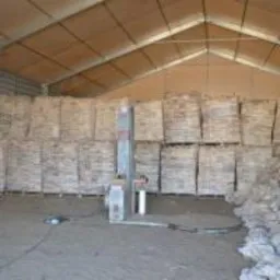 Magazyn z zapasem drewna opałowego w workach, zabezpieczonego folią. W tle widoczne ściany magazynu i metalowa konstrukcja podtrzymująca dach. Częściowo widoczna maszyna do pakowania.