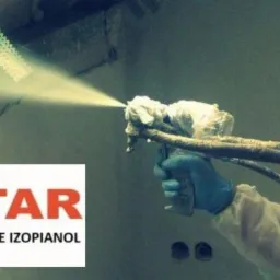 Aplikacja izolacji natryskowej: pracownik w kombinezonie ochronnym nanosi pianę na poddasze, widoczne metalowe elementy konstrukcyjne i świeżo nałożona warstwa izolacji.