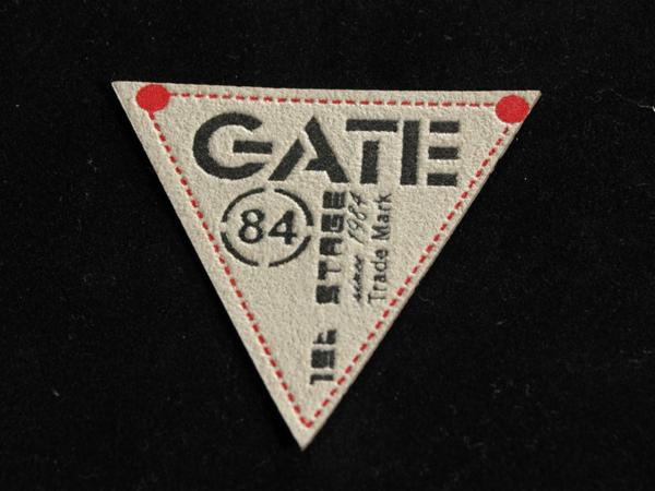 Trójkątna metka z logo 'GATE 84', czerwoną obwódką i dwoma czerwonymi punktami w rogach, umieszczona na czarnym tle. Detal wykończenia odzieży.