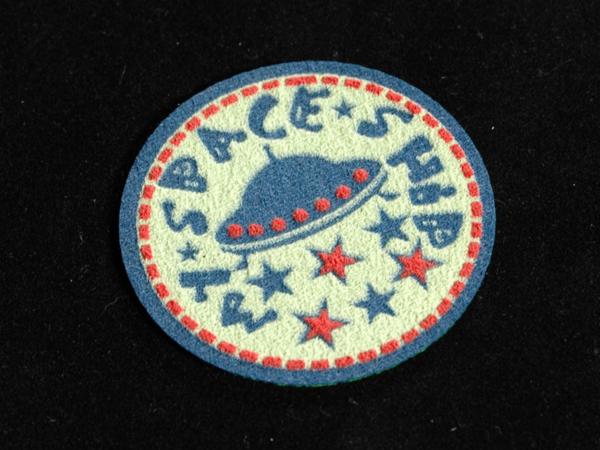 Okrągła naszywka z motywem UFO i gwiazd na czarnym tle. Stylizowane litery tworzą napis 'SPACE SHIP'. Drobne przeszycia na obrzeżach.