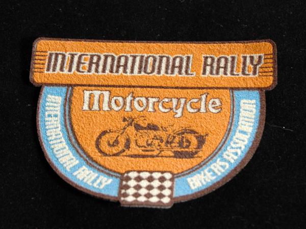 Naszywka z napisem 'International Rally Motorcycle Bikers Association' i rysunkiem motocykla, utrzymana w odcieniach brązu i niebieskiego, na czarnym tle.