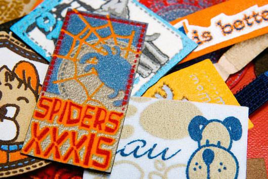 Kolekcja tkanych naszywek z różnymi wzorami: pająk, pieski, napisy. Naszywki o zróżnicowanej kolorystyce i fakturze, ułożone blisko siebie. Detal nadruku na tkaninie.