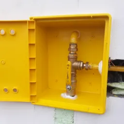 Żółta skrzynka gazowa z zaworem i rurami na białej ścianie z widoczną pianką montażową.