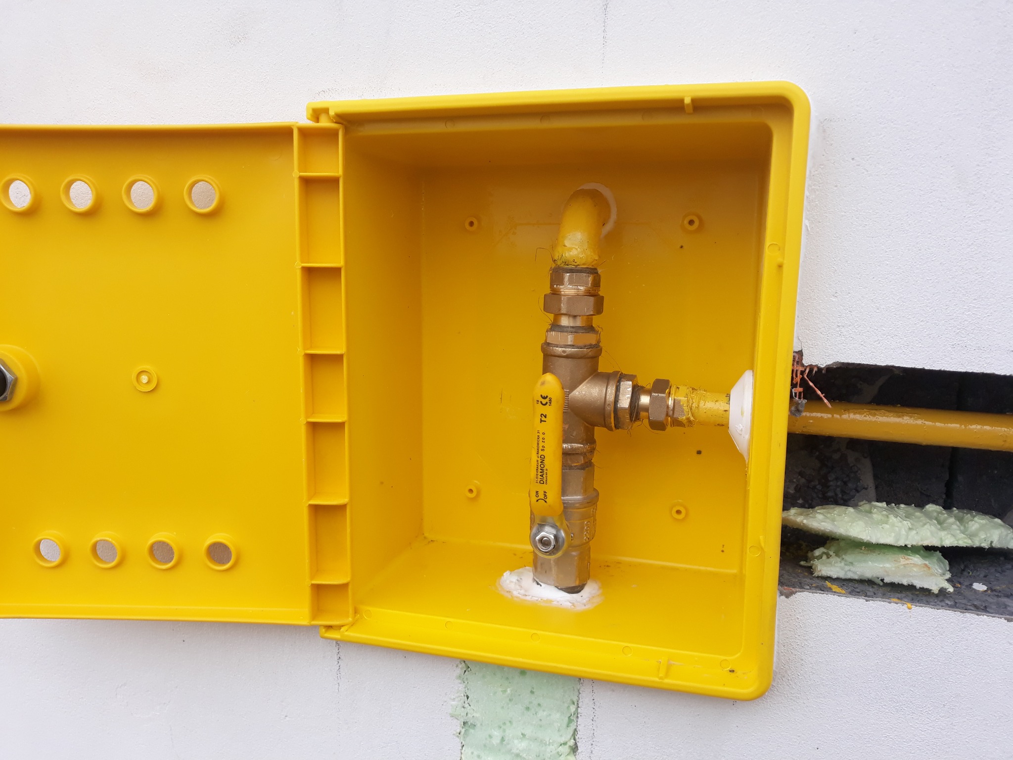 Żółta skrzynka gazowa z zaworem i rurami na białej ścianie z widoczną pianką montażową.