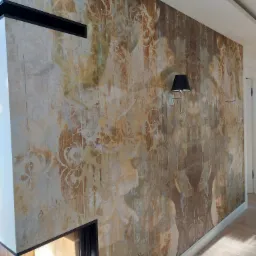 Ściana z dekoracyjną tapetą w odcieniach brązu i beżu, oświetlona kinkietem z czarnym abażurem, obok nowoczesnego kominka z czarną obudową.