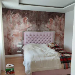 Sypialnia z łóżkiem z pikowanym, różowym zagłówkiem i wzorzystą tapetą w odcieniach brązu i czerwieni. Na podłodze drewniany parkiet, obok łóżka stos książek.